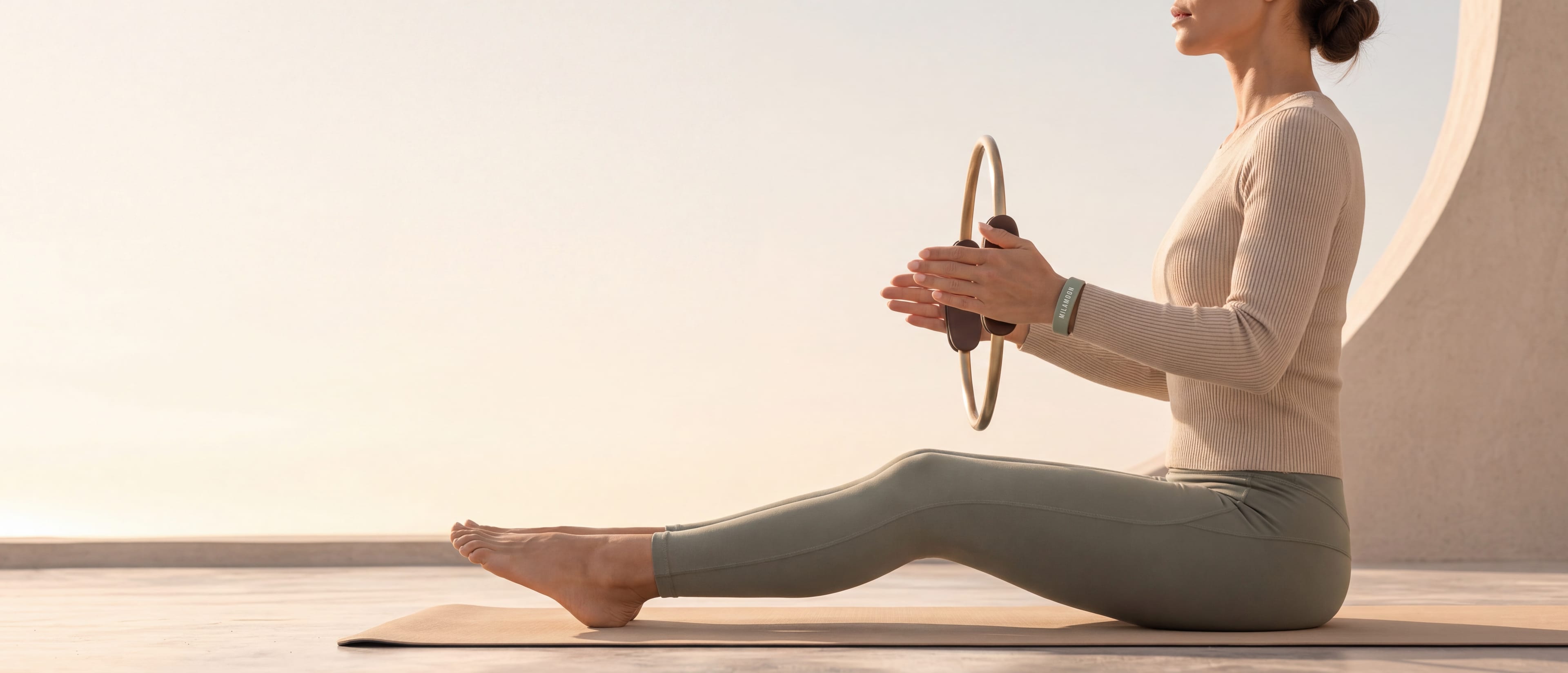 A woman sits in profile on a mat, pressing a Pilates ring between her hands, the MILA MOON wristband on her wrist