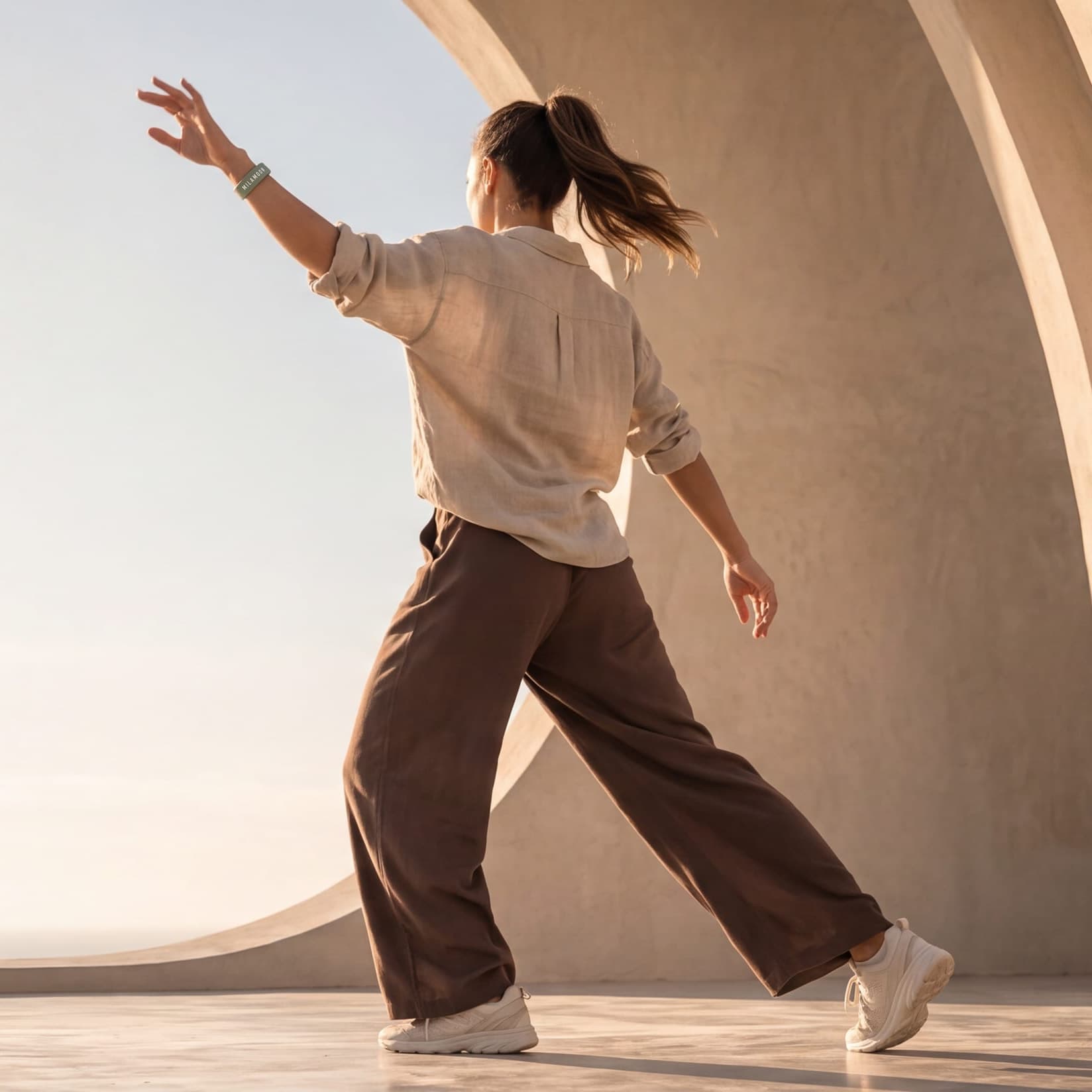 A woman mid dance step in front of curved walls, arm raised, the MILA MOON wristband on her wrist