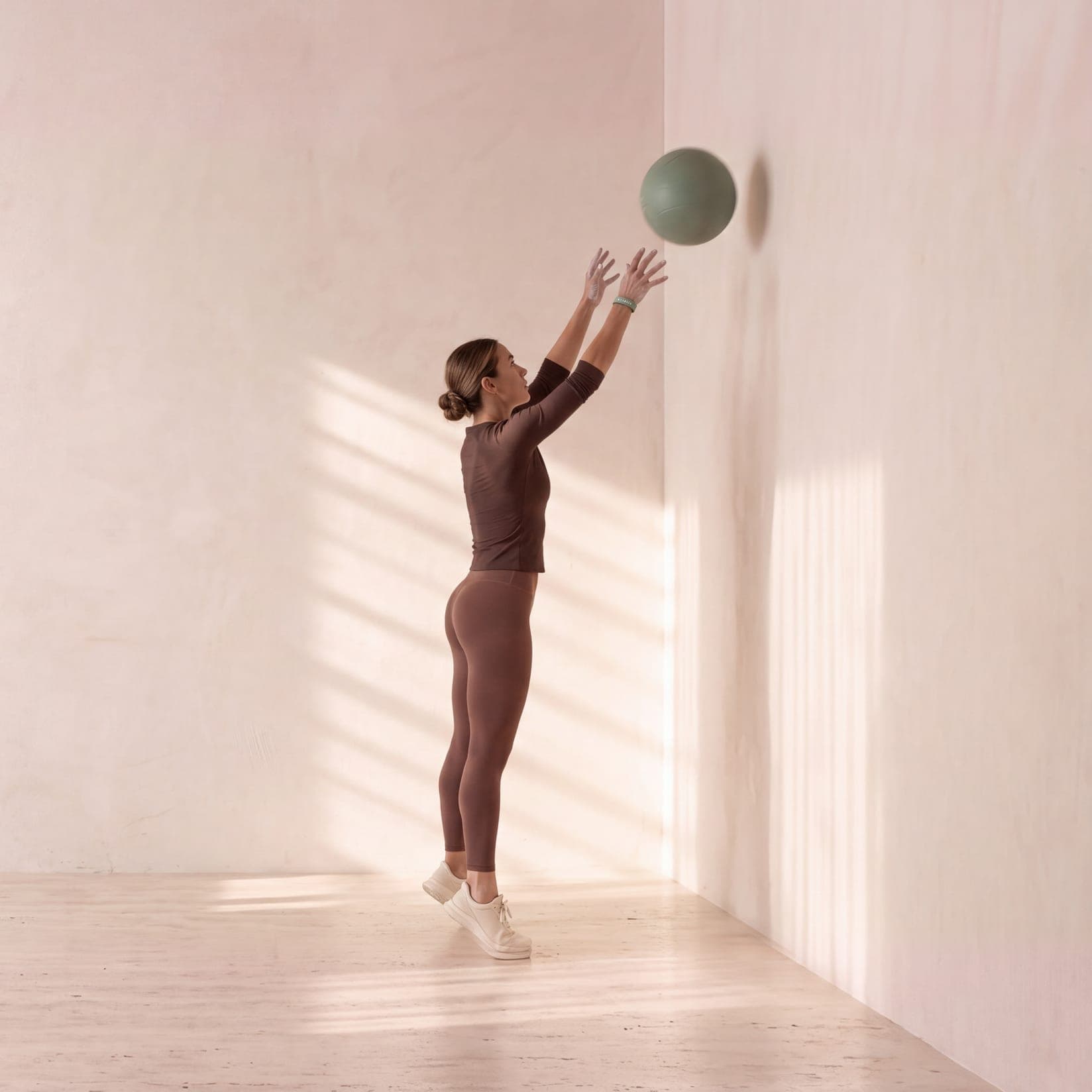 Eine Frau mit dem MILA-MOON-Armband wirft auf den Zehenspitzen einen salbeigrünen Medizinball an die Wand
