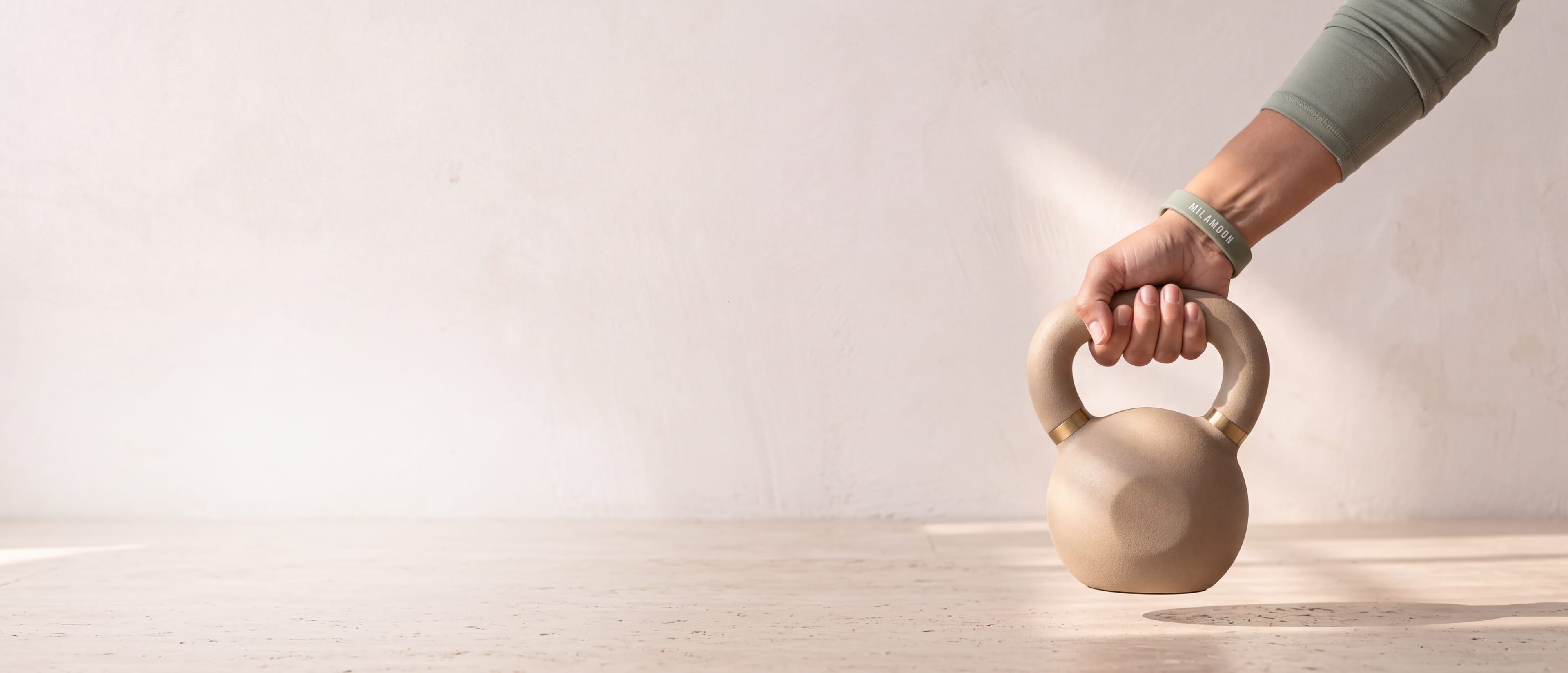 Eine Hand mit dem MILA-MOON-Armband hebt eine sandfarbene Kettlebell vom hellen Steinboden