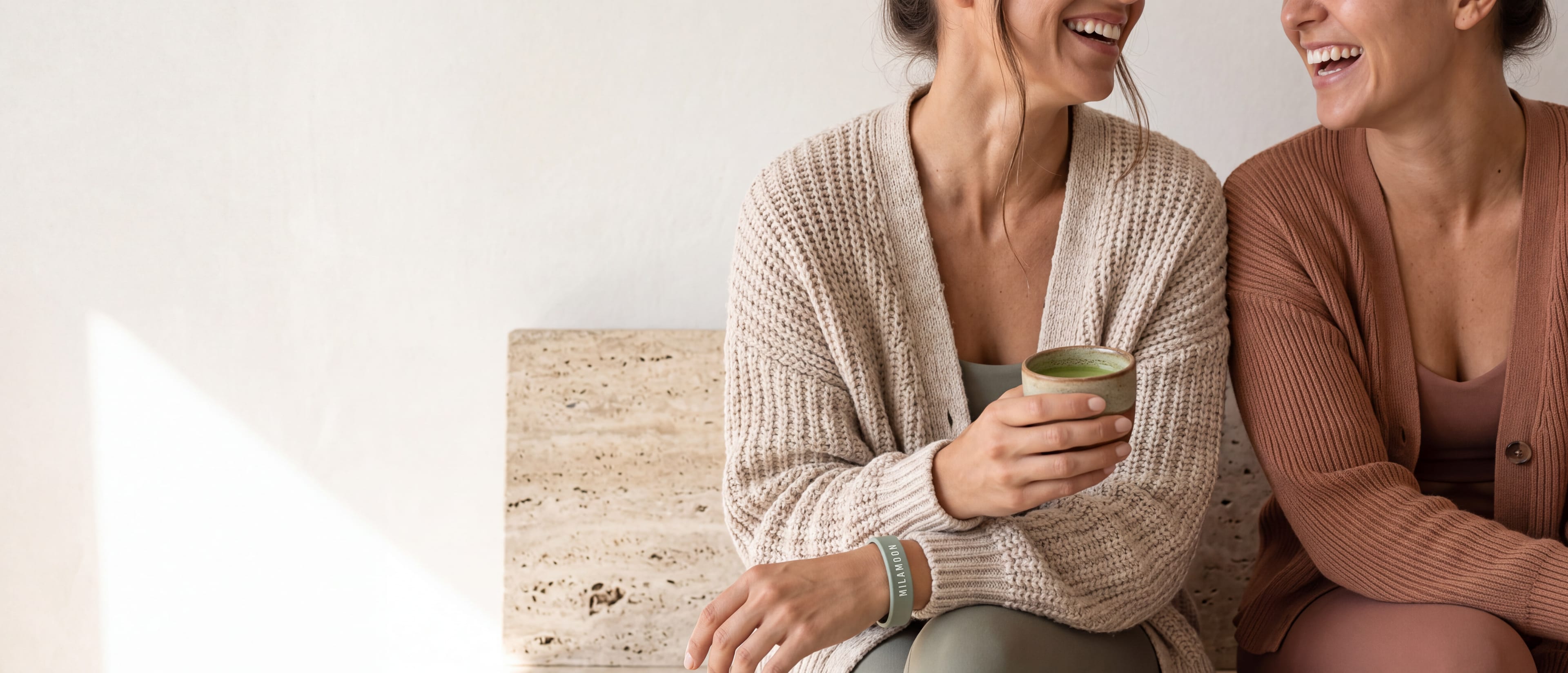 Zwei Frauen in Strickjacken lachen nach dem Training, eine hält einen Matcha und trägt das MILA-MOON-Armband